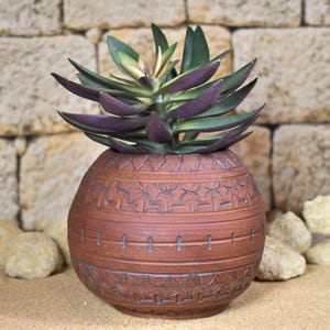 May include: A decorative, round, terracotta-colored ceramic pot with etched geometric patterns, holding a succulent plant with green and purple leaves. The pot sits on sand with small rocks, against a stone wall background.