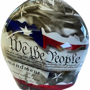 May include: A glass paperweight featuring a printed design of the United States Constitution with the text We the People and Amendment. The surface displays red, white, and blue flag motifs, white stars, and black script text on a parchment-coloured background.