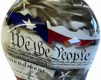 May include: A glass paperweight with a red, white, and blue design. The paperweight features a stylized image of the Texas flag and the text "We the People" from the US Constitution.