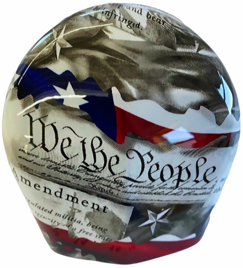 May include: A glass paperweight featuring a printed design of the United States Constitution with the text We the People and Amendment. The surface displays red, white, and blue flag motifs, white stars, and black script text on a parchment-coloured background.