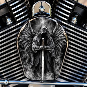May include: A motorcycle engine cover featuring a black and white graphic of a hooded figure with large feathered wings, holding a long sword upright. The figure is surrounded by swirling smoke, set against a background of metallic engine fins.