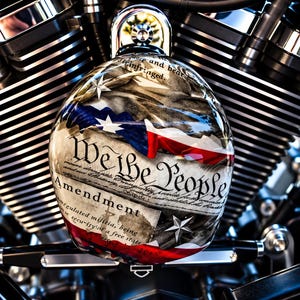 May include: A round, glossy motorcycle air cleaner cover featuring a graphic of the United States Constitution with the text We the People and Amendment, overlaid with a stylised American flag design and silver stars, mounted on a black engine.