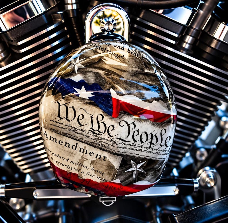 May include: A round, glossy motorcycle air cleaner cover featuring a graphic of the United States Constitution with the text We the People and Amendment, overlaid with a stylised American flag design and silver stars, mounted on a black engine.