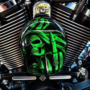 May include: A motorcycle air cleaner cover featuring a vibrant green airbrushed graphic of a hooded skull making a middle finger gesture. The glossy, oval-shaped cover is mounted on a black engine block with silver cooling fins and a Harley-Davidson logo.