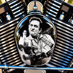 May include: A black and white circular motorcycle air cleaner cover featuring a portrait of Johnny Cash holding an acoustic guitar and gesturing with a middle finger. The cover is mounted against a background of chrome engine cooling fins.