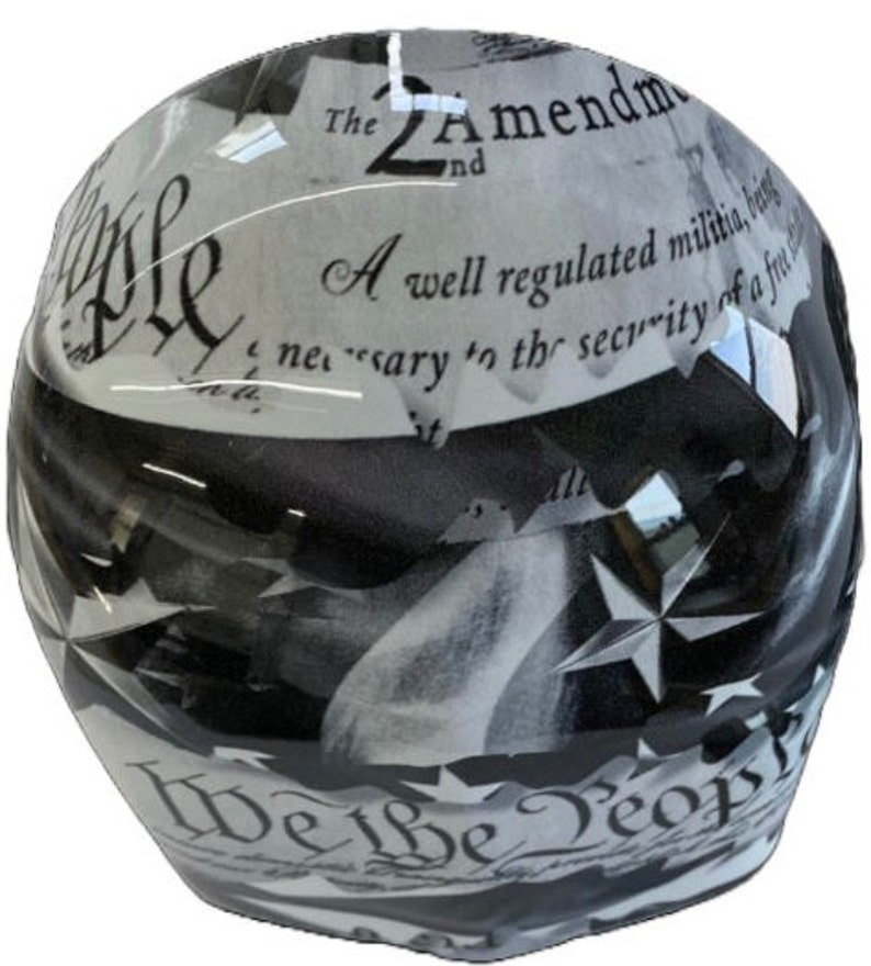 May include: A glossy motorcycle helmet featuring a black and white graphic design. The surface displays text from the 2nd Amendment and the phrase We the People in a decorative script font, interspersed with star shapes across the rounded shell.
