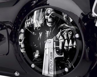 May include: A circular black motorcycle engine derby cover featuring a monochromatic graphic of a skeleton wearing a jacket and spiked gloves, holding a handgun with the text BAD ASS engraved on the side of the firearm barrel.