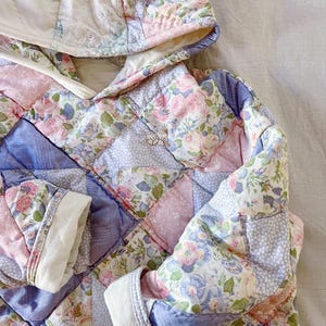 Könnte beinhalten: Eine gesteppte Jacke mit einem Blumendruck in Rosa-, Blau- und Weißtönen. Die Jacke hat eine Kapuze und lange Ärmel.