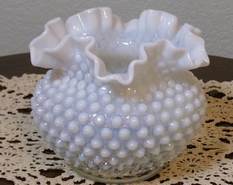 May include: A small, round, light blue opalescent glass vase featuring a hobnail texture across its exterior surface. The vase has a wide, ruffled, and crimped rim that flares outward, creating a decorative, wavy edge around the opening.