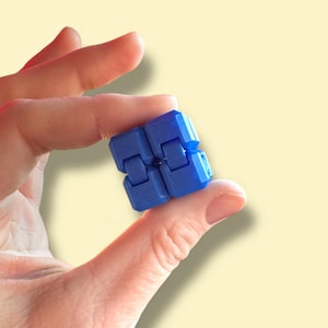 May include: A small, blue fidget toy held between fingers. The toy is a cube-like shape, made of connected blocks that can be manipulated. The background is a soft yellow.