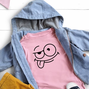 Peut inclure: Un t-shirt rose avec un visage de dessin anim&eacute; noir avec la langue qui sort. Le t-shirt est superpos&eacute; &agrave; une veste en jean bleu avec une capuche.
