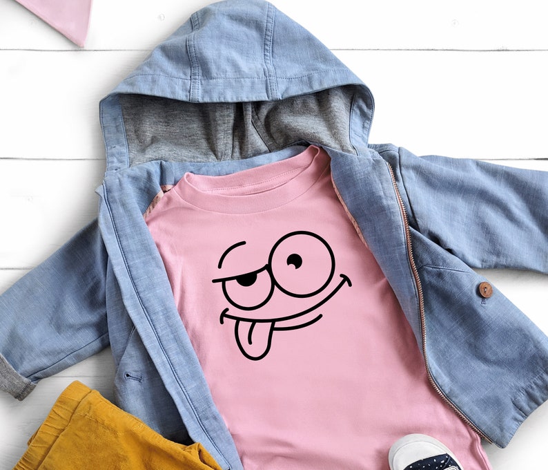 Peut inclure: Un t-shirt rose avec un visage de dessin anim&eacute; noir avec la langue qui sort. Le t-shirt est superpos&eacute; &agrave; une veste en jean bleu avec une capuche.