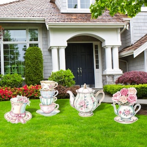May include: Lawn decorations featuring a whimsical tea party theme. Includes a stack of teacups, a teapot, a hat, and a teapot filled with roses. The decorations are set on a green lawn in front of a house.