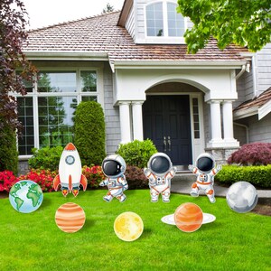 May include: A set of space-themed yard signs featuring a white and orange rocket, three astronauts in white suits, and five planetary bodies including Earth, a yellow moon, and ringed planets, all displayed on a green lawn in front of a house.