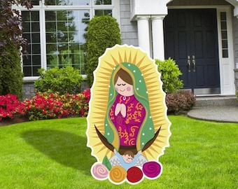 May include: A colorful, cartoon-style cutout of the Virgin Mary with a halo and a green and pink robe. The cutout is standing on a lawn with five pink, orange, and red roses at her feet.
