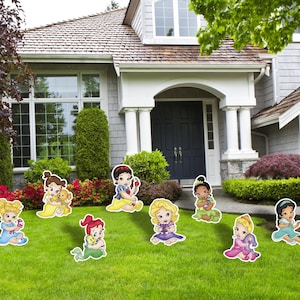May include: A collection of eight colorful cartoon character lawn ornaments featuring stylized infants in various outfits, including blue, yellow, red, purple, green, and pink dresses, arranged across a green grass lawn in front of a house.