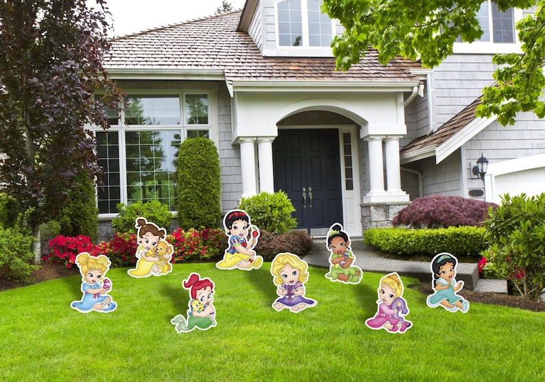May include: A collection of eight colorful cartoon character lawn ornaments featuring stylized infants in various outfits, including blue, yellow, red, purple, green, and pink dresses, arranged across a green grass lawn in front of a house.