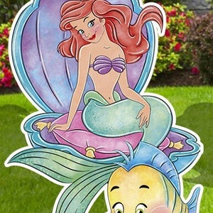 Puede incluir: Un recorte de Ariel, la Sirenita, sentada en una concha p&uacute;rpura con un coj&iacute;n rosa. Lleva un sujetador de concha p&uacute;rpura y una cola verde. Un pez amarillo con aletas azules est&aacute; debajo de ella.