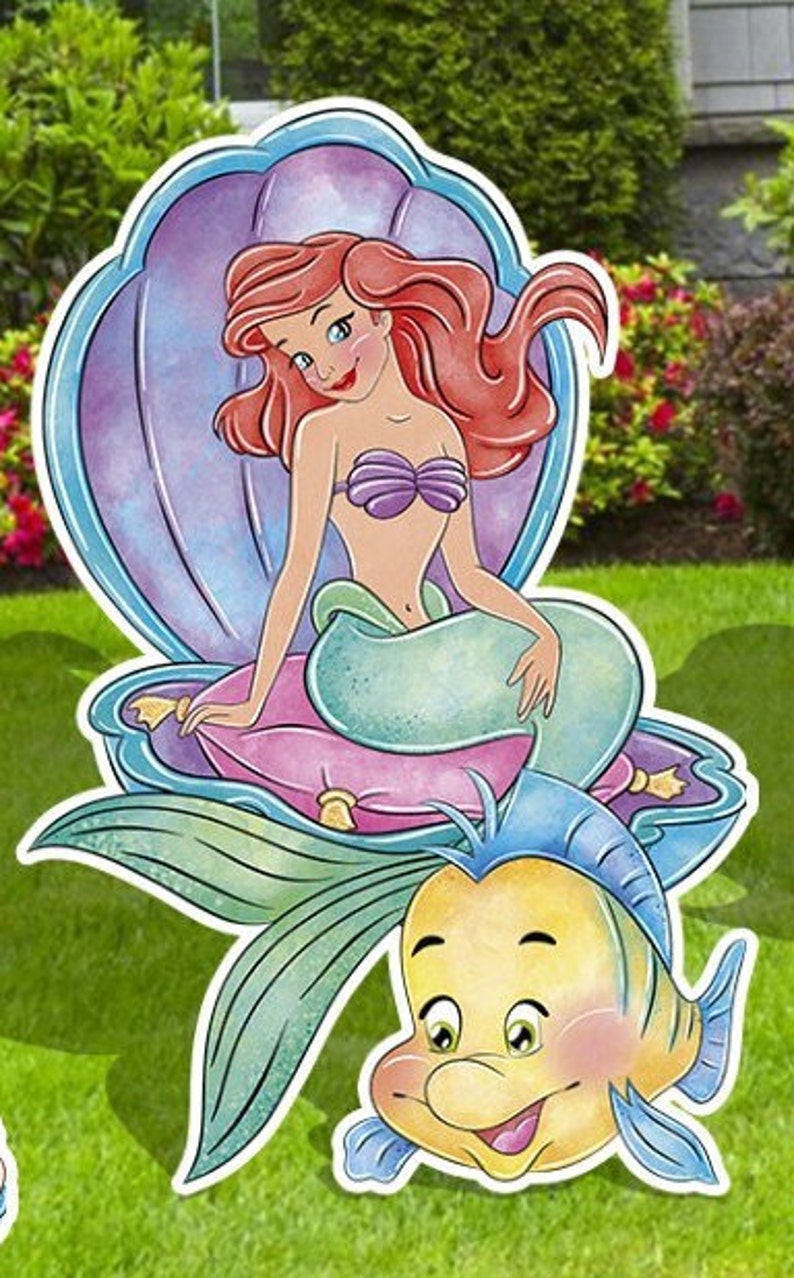 Puede incluir: Un recorte de Ariel, la Sirenita, sentada en una concha p&uacute;rpura con un coj&iacute;n rosa. Lleva un sujetador de concha p&uacute;rpura y una cola verde. Un pez amarillo con aletas azules est&aacute; debajo de ella.