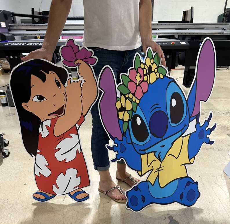 May include: Two large cardboard cutouts of cartoon characters. The character on the left is a girl with long black hair, wearing a red and white dress. The character on the right is a blue alien with large ears, wearing a yellow shirt and a flower crown.