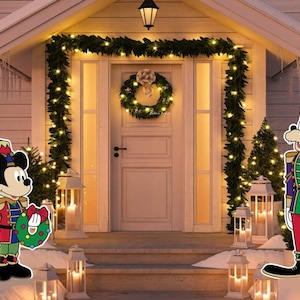 May include: A festive Christmas scene with a Mickey Mouse and Goofy cutout. Mickey is dressed as a toy soldier holding a wreath. Goofy is dressed as a toy soldier holding a candy cane. Both are standing in front of a house decorated with Christmas lights and greenery.