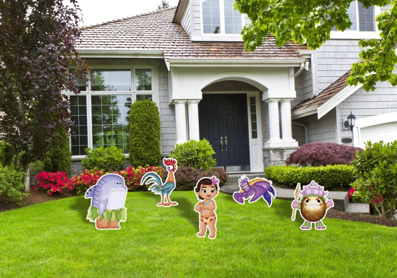 May include: A collection of colorful character cutouts, including a shark, rooster, girl, crab, and coconut, are displayed on a green lawn in front of a house. The cutouts are designed with cartoon-like features and vibrant colors.