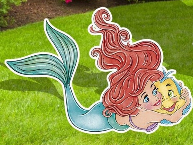 Puede incluir: Un recorte de Ariel, la Sirenita, con cabello rojo y cola azul, abrazando a un Flounder amarillo. El recorte est&aacute; sobre un fondo de hierba verde.