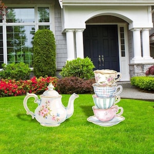 May include: A decorative outdoor set featuring a teapot and a stack of teacups. The teapot is white with pink floral accents. The teacups have various patterns, including floral, plaid, and striped designs. The set is placed on a green lawn.