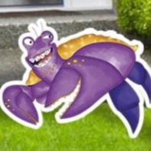 May include: A whimsical, cartoon-style purple hermit crab yard decoration with a yellow shell and a big, toothy grin. The crab has large claws and is set against a backdrop of green grass and a concrete surface.