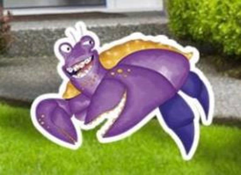 May include: A whimsical, cartoon-style purple hermit crab yard decoration with a yellow shell and a big, toothy grin. The crab has large claws and is set against a backdrop of green grass and a concrete surface.