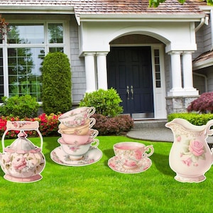 May include: A set of pink and white teacups, teapot, and creamer, decorated with floral designs. The teacups are stacked on top of each other, and the teapot and creamer are sitting on the ground. The set is in front of a house with a green lawn.