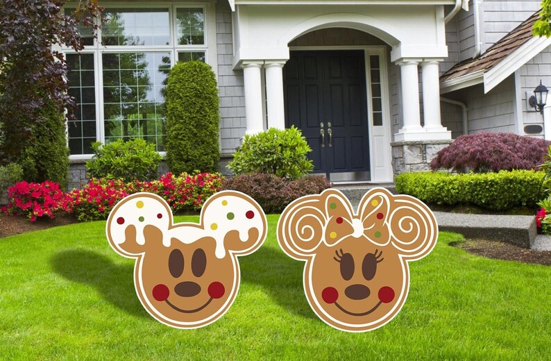 Puede incluir: Dos decoraciones de jard&iacute;n con tem&aacute;tica de pan de jengibre con Mickey Mouse y Minnie Mouse. Las decoraciones son marrones con glaseado blanco y chispas de colores. Mickey Mouse lleva una pajarita roja y Minnie Mouse lleva un lazo rojo.
