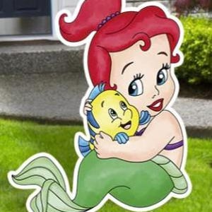 May include: A decorative yard sign featuring a cartoon illustration of a red-haired mermaid with a green tail, embracing a small yellow and blue tropical fish. The cutout has a white border and depicts the characters with large eyes and smiles.