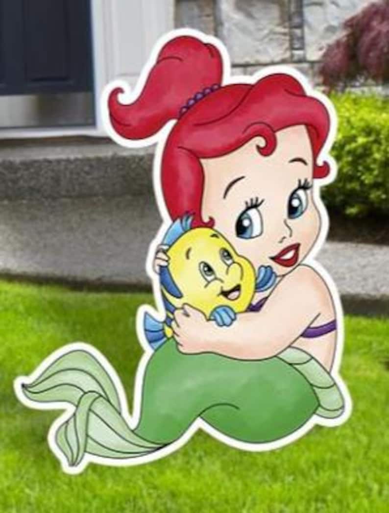 May include: A decorative yard sign featuring a cartoon illustration of a red-haired mermaid with a green tail, embracing a small yellow and blue tropical fish. The cutout has a white border and depicts the characters with large eyes and smiles.