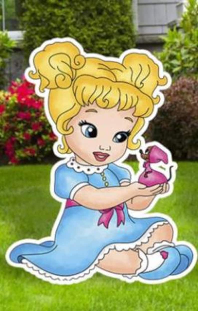 May include: A cartoon-style cutout of a young child with blonde hair in two buns, wearing a light blue dress with a pink sash and white trim. The child is holding a small pink mouse figure in their hands. The illustration features bold outlines.