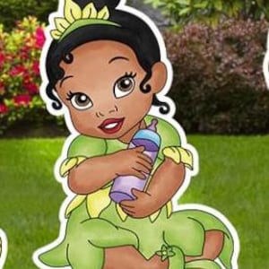 May include: A cartoon illustration of a baby with dark skin and black hair styled in a bun, wearing a green dress with yellow petal accents and a matching headband. The baby is holding a purple baby bottle with a light blue cap.