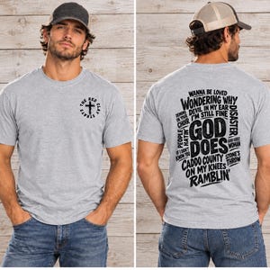 Peut inclure: Un t-shirt gris clair à manches courtes avec un petit logo circulaire sur la poitrine représentant une croix et le texte The Red Clay Strays. Le dos présente un grand texte noir incluant les mots God Does et diverses phrases.