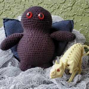 Puede incluir: Un monstruo de amigurumi de crochet negro con ojos rojos se sienta en una manta de crochet blanca con un juguete de esqueleto de plástico blanco.