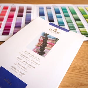 May include: Image of DMC embroidery floss colour charts and product brochures. The charts display a wide range of colours, from pinks and reds to blues and greens. The brochures feature product names and the DMC logo.