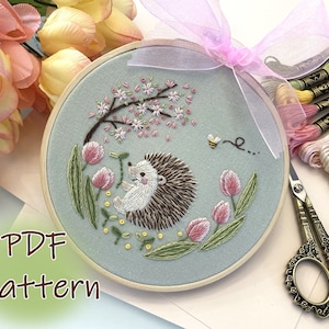 May include: Embroidered hoop art featuring a hedgehog sitting under a blooming cherry blossom tree with a bee flying nearby. The hoop is on a white surface with pink and yellow flowers and embroidery floss.
