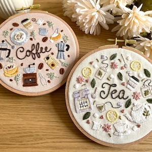 Peut inclure: Deux cerceaux à broder avec un tissu beige et des cadres en bois brun. Le cerceau de gauche présente un thème café avec des images brodées de grains de café, un moulin à café, une cafetière et le mot "Café". Le cerceau de droite présente un thème thé avec des images brodées de tasses à thé, de feuilles de thé, une théière et le mot "Thé".