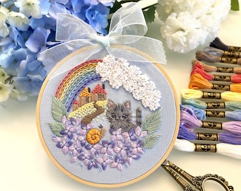May include: Embroidered hoop art featuring a black cat, a rainbow, a snail, and flowers on a light blue background. The hoop is surrounded by a white ribbon.