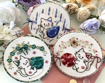May include: Three embroidery hoops with white felt backgrounds and colorful embroidery designs. Each hoop features a white cat with a different colored gemstone. The first hoop has a blue gemstone and blue flowers, the second hoop has a green gemstone and green leaves, and the third hoop has a red gemstone and pink flowers.