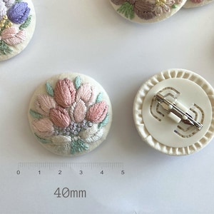 Puede incluir: Un broche de tela blanca con un dise&ntilde;o de flor bordada en rosa y verde. La parte posterior del broche tiene un cierre de metal blanco. El broche mide 40 mm de di&aacute;metro.