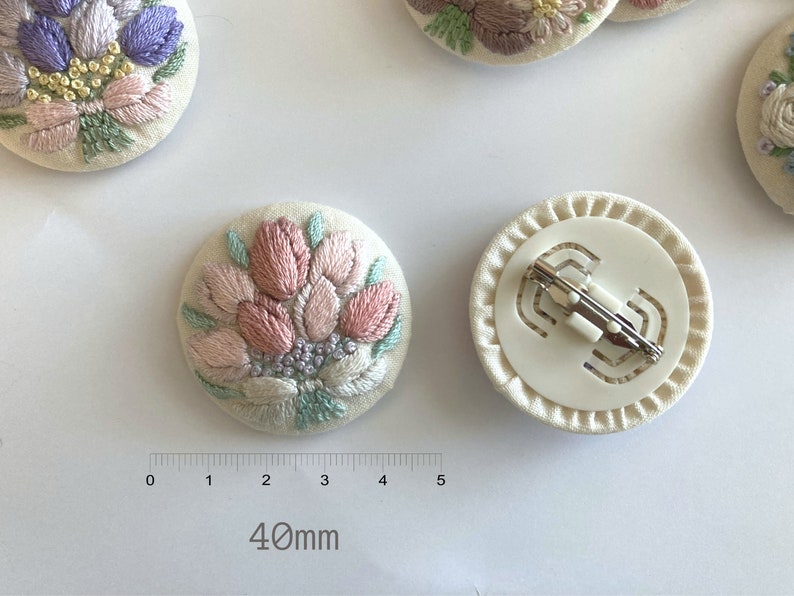 Puede incluir: Un broche de tela blanca con un dise&ntilde;o de flor bordada en rosa y verde. La parte posterior del broche tiene un cierre de metal blanco. El broche mide 40 mm de di&aacute;metro.