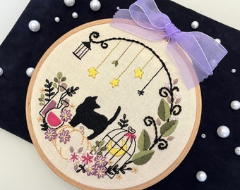 May include: A white embroidery hoop featuring a black cat, a birdcage, and floral designs. The hoop is adorned with a purple ribbon.