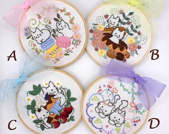 May include: Four circular wooden embroidery hoops featuring white fabric with stitched cat designs. Hoop A shows cats with macarons, B with pastries, C with cupcakes and strawberries, and D with candy. Each hoop is topped with a sheer colored ribbon.