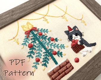 May include: A cross-stitch embroidery pattern featuring a cat wearing a Santa hat holding a basket of red ornaments in front of a Christmas tree with red ornaments and a string of lights. The text "PDF Pattern" is in the lower left corner of the image.