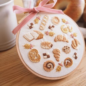 May include: A circular wooden embroidery hoop containing white fabric with detailed thread-stitched patterns of various baked goods including a croissant, pretzel, baguette, and pastries, accented by a soft pink velvet ribbon tied at the top.
