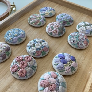 Puede incluir: Diez broches florales bordadas en varios colores y dise&ntilde;os, expuestas sobre una superficie de madera.